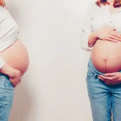Dwa zaokrąglone brzuchy w 4 miesiącu ciąży, obie kobiety w białych koszulach i jeansach.