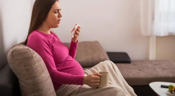 Ciężarna kobieta z kubkiem w dłoni i chusteczką, szukająca informacji co na kaszel w ciąży.