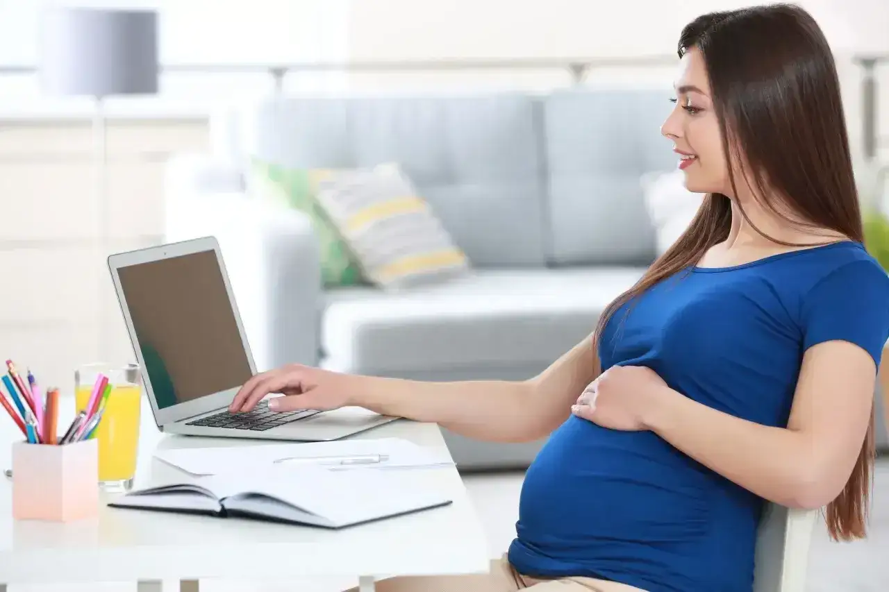 Uśmiechnięta kobieta w zaawansowanej ciąży, w niebieskiej sukience, pracuje przy laptopie. W tle salon. L4 ciążowe.