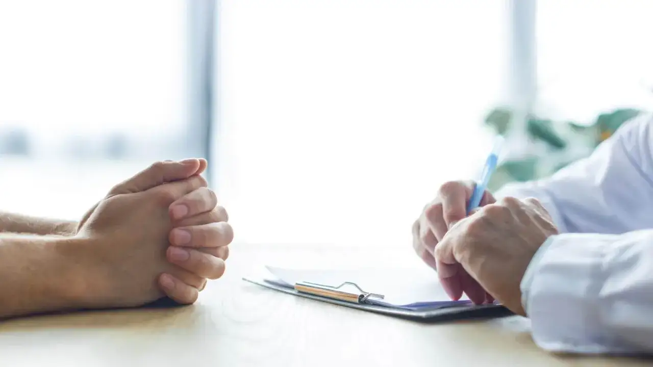 Pacjent z zaciśniętymi dłońmi słucha lekarza, który pisze na dokumentach. Sytuacja może dotyczyć wypowiedzenia umowy o pracę a zwolnienia lekarskiego od psychiatry.