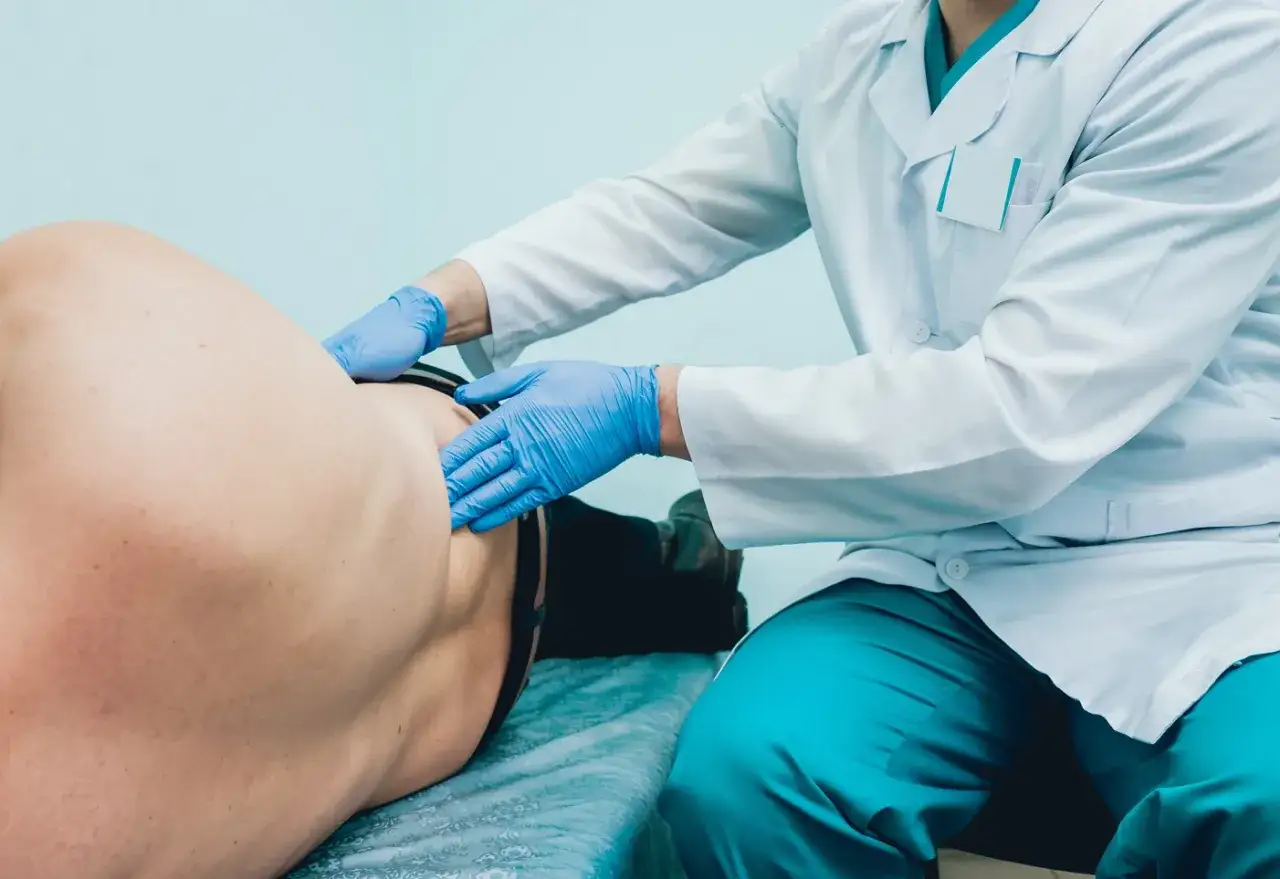 Urolog bada pacjenta. Lekarz w białym fartuchu i niebieskich rękawiczkach delikatnie uciska plecy pacjenta, aby ocenić jego stan.