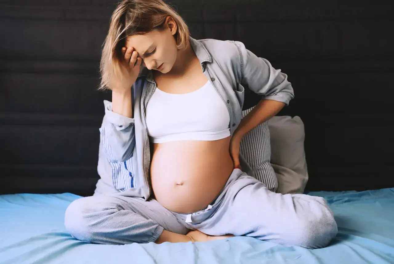 Ciężarna kobieta z bólem krocza w ciąży, trzymająca się za głowę i plecy, siedzi na łóżku.